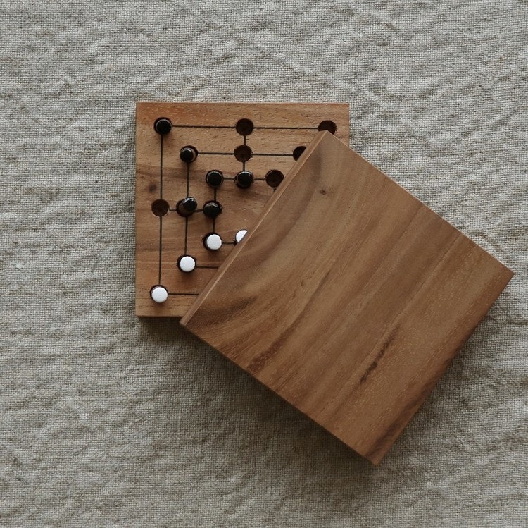A small, handmade board game made from teak. Square, 11x11 cm with lid and black and white wooden pegs.