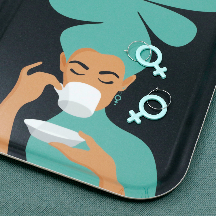 Detaljbild på en turkos bricka med motivet Kaffekvinna. Kaffekvinnan har stort böljande hår, bär ett feministörhänge och njuter av en kopp kaffe. På brickan ligger två mintfärgade feministörhängen.