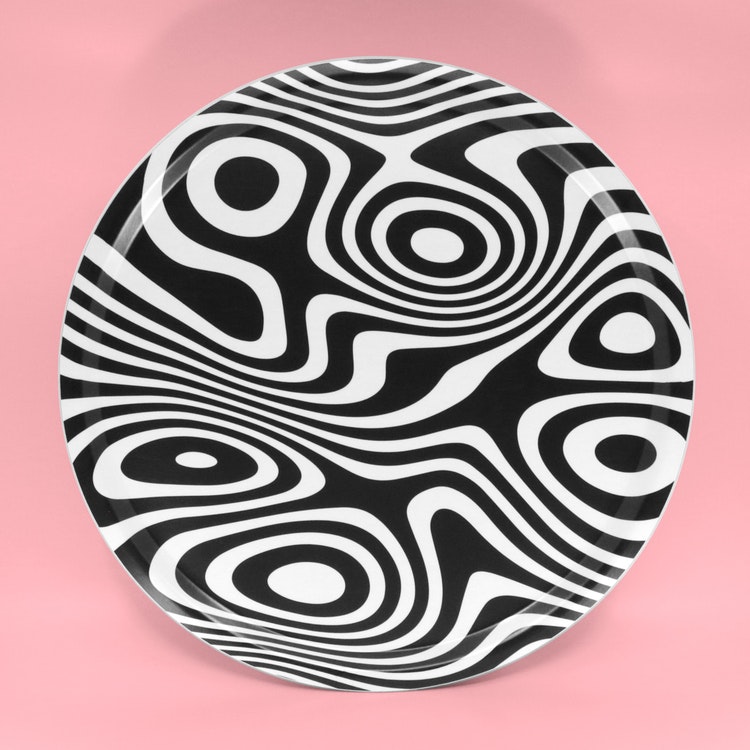 Rund svartvit bricka med ett abstrakt, snurrigt och hypnotiskt mönster.
