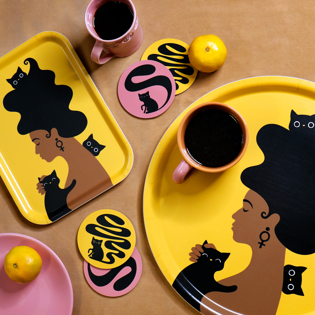 Två gula brickor – en rektangulär och en rund – med motivet Kattkvinnan – en kvinna med stort böljande hår, en feministsymbol / venussymbol /kvinnosymbol i örat, omgiven av tre busiga katter. Med på b