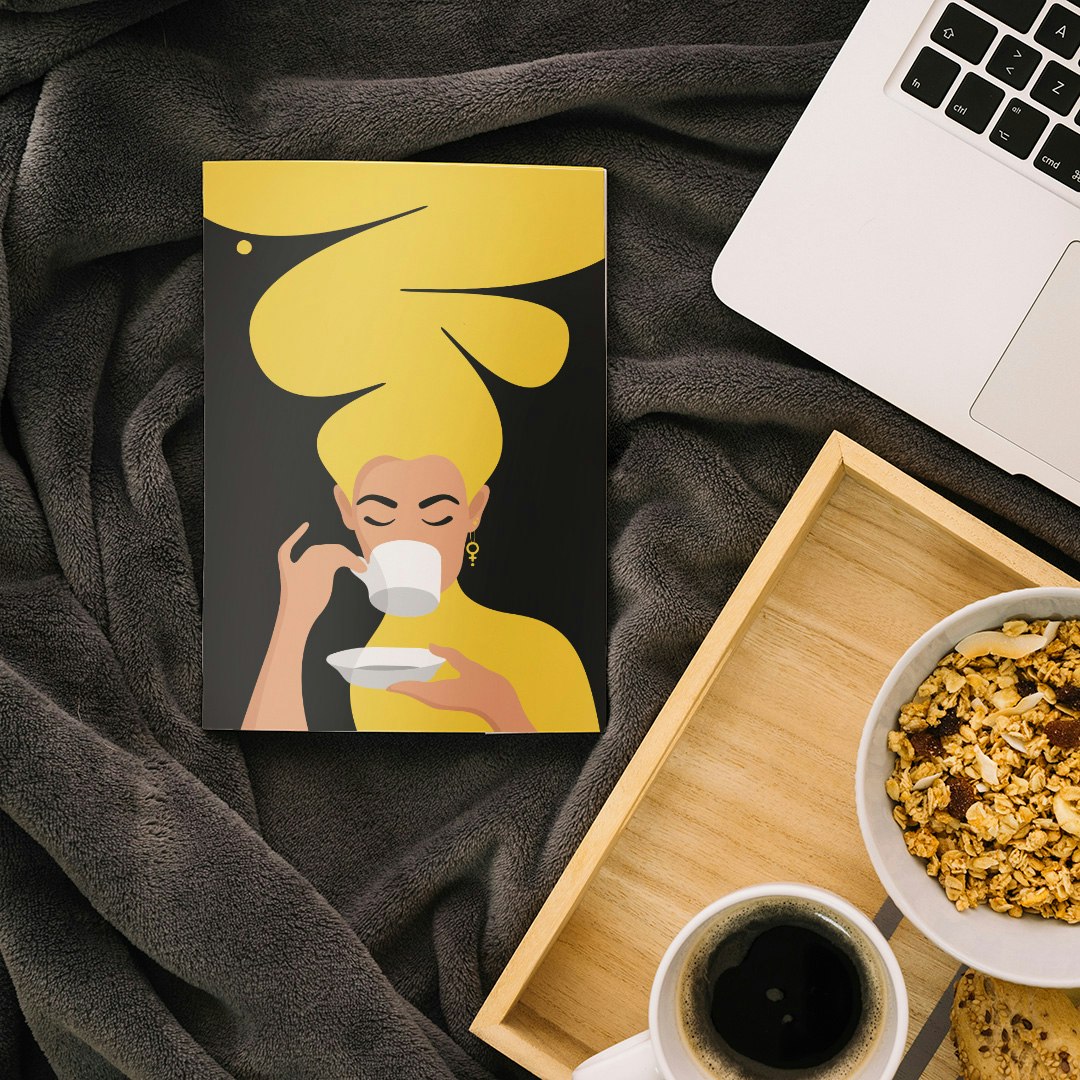 Ritblock / skissblock / anteckningsblock med motivet Kaffekvinnan i gult på omslaget. Liggandes bredvid frukost och laptop.