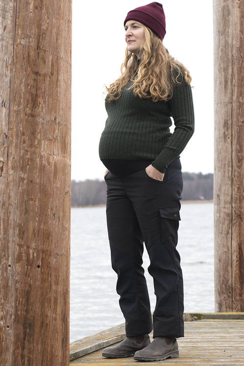 En gravid kvinna i ett par svarta servicebyxor står på en brygga och tittar ut över vattnet.