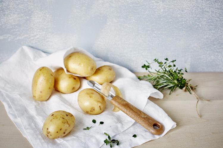 Naturfärgad potatisskalare i rostfritt stål med skaft av bioplast och träfiber på en vit duk med potatis brevid.