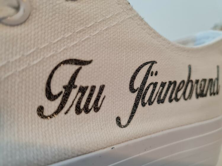 Bild av en sko i textil med en text i textilvinyl, där kunden kan beställa med egen text. Anpassningsbar i färgerna vit, svart, guld och silver.