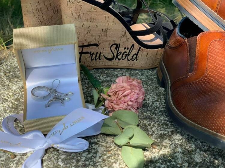 Bild av bröllopsaccessoarer utomhus: ett par bruna läderskor, en matchande läderbälte, en träask märkt 'Fru Sköld', en ringask med förlovningsring och en rosa blomma. Detaljerade och eleganta föremål 
