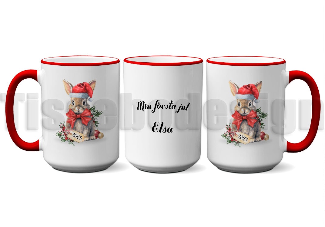 Vit mugg med rött handtag och kant, motiv av en kanin i tomteluva och texten "Min Första Jul" med namnet Elsa, porslin 450 ml, tryck på båda sidor.