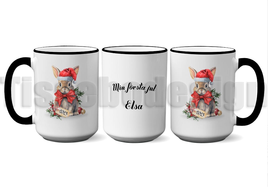 Vit mugg med svart handtag och kant, motiv av en kanin i tomteluva och texten "Min Första Jul" med namnet Elsa, porslin 450 ml, tryck på båda sidor.