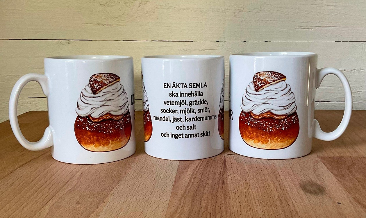 Vit keramikmugg med texten En äkta semla ska innehålla vetemjöl, grädde, socker, mjölk, smör, mandel, jäst, kardemumma och salt och semmelmotiv på båda sidor, 330 ml