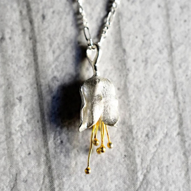 Bild på ett silverhalsband med ett hänge format som en blomklocka med guldpläterade detaljer.