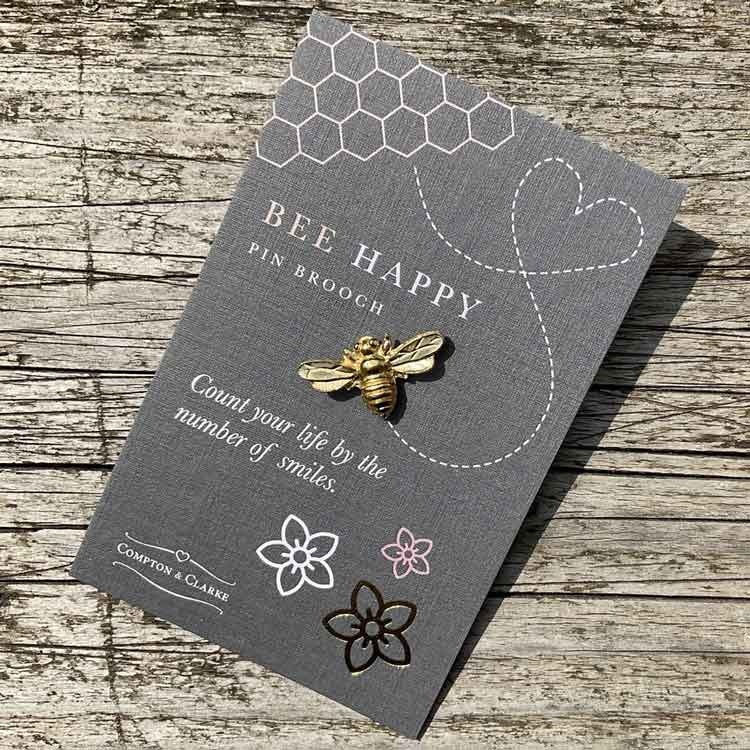 Bild på en pin i form av ett bi tillverkat i tenn som guldpläterats. Pinen är fäst på ett litet kort med texten "bee happy"