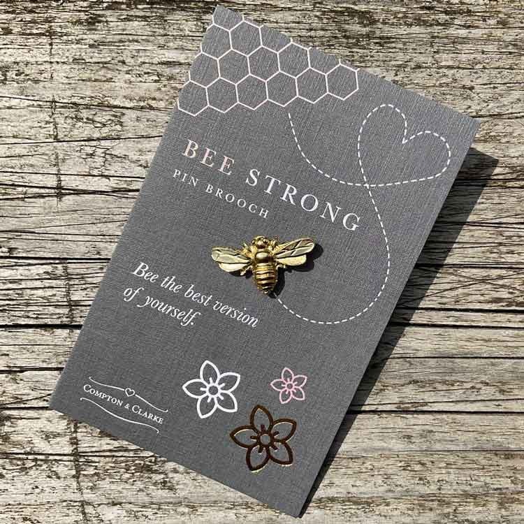 Bild på en pin i form av ett bi tillverkat i tenn som guldpläterats. Pinen är fäst på ett litet kort med texten "bee strong"