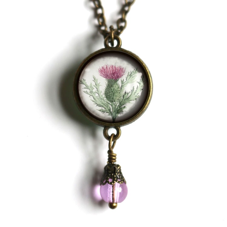 Bild på ett vintageinspirerat halsband med en illustration av en tistel samt en pärla under.