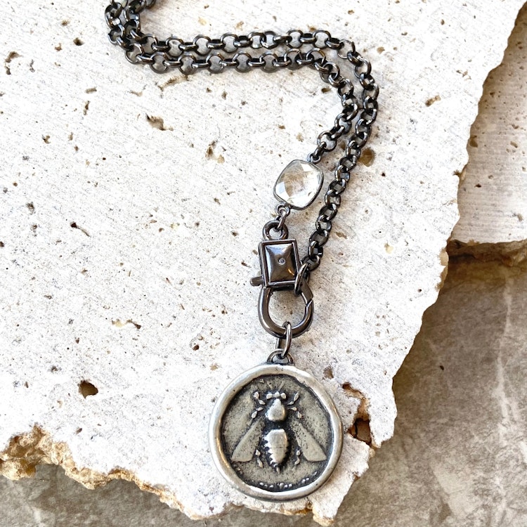 Bilden visar ett halsband med en grov kedja med en kristall samt ett hönge med ett bi.