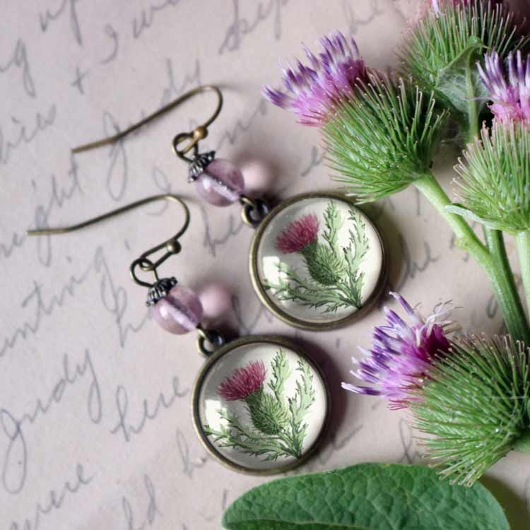 Örhängen med pärlor och illustrationer av tistlar under cabochoner av glas.
