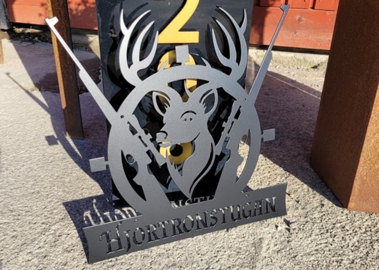 Metallskylt med utskuret namn och motiv av en hjort och korsade gevär.