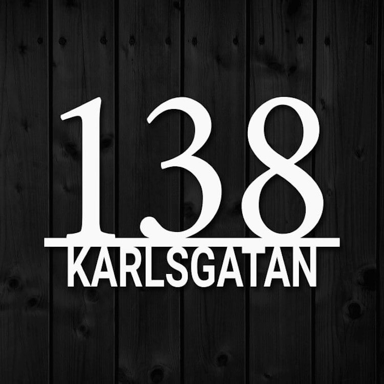 Vit husnummerskylt i metall, mot svart träfasad.