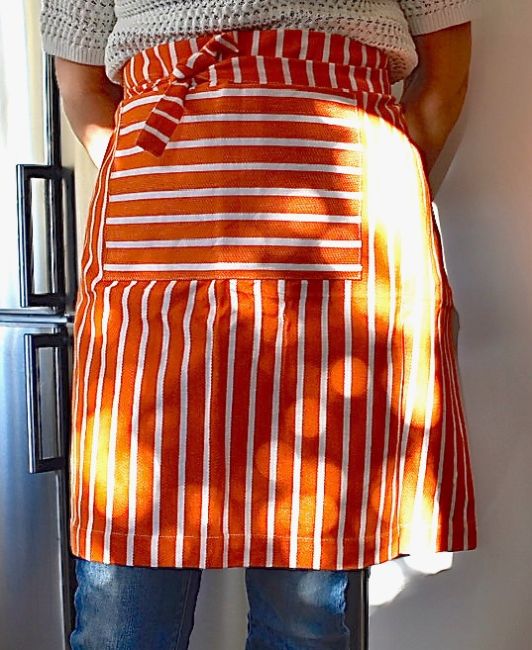 Poppy ett orange och vitrandigt förkläde i 100% bomull från Noble house, mått 60 x 75 cm.