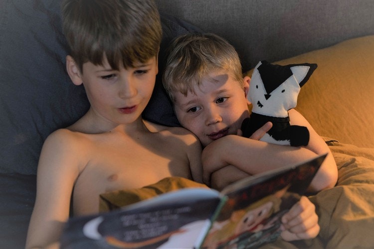 Två barn läser godnattsaga i en säng. Det ena barnet håller en djurkompis, tygdjur, mjukisdjur  i famnen.