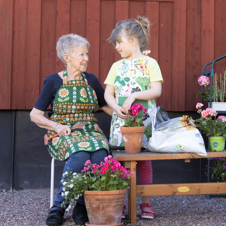 Mormor, farmor, pensionär, äldre kvinna med ett barn, barnbarn. Båda har på sig praktiska trädgårdsförkläden när de håller på att plantera om blommor.