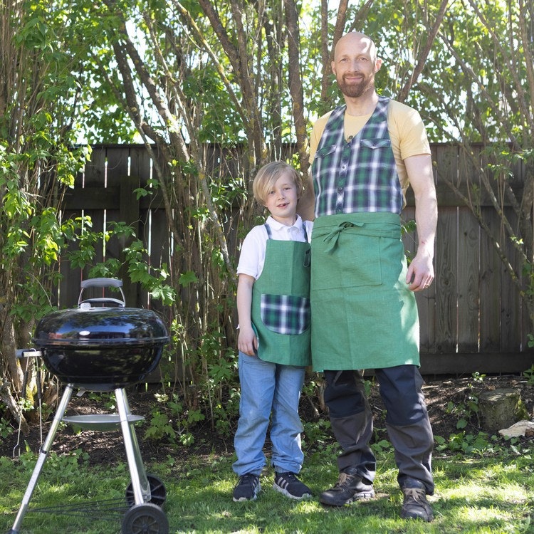 Far och son vid grillen i trädgården. Pappa har ett förkläde "Köksan" av rutig skjorta i grönt och blått samt grönt linne. Barnet har ett förkläde i samma material.