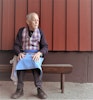 En ensam äldre man på en bänk. Iklädd förkläde "Köksan" skapat av minnestextilier från nära bortgången som han saknar.