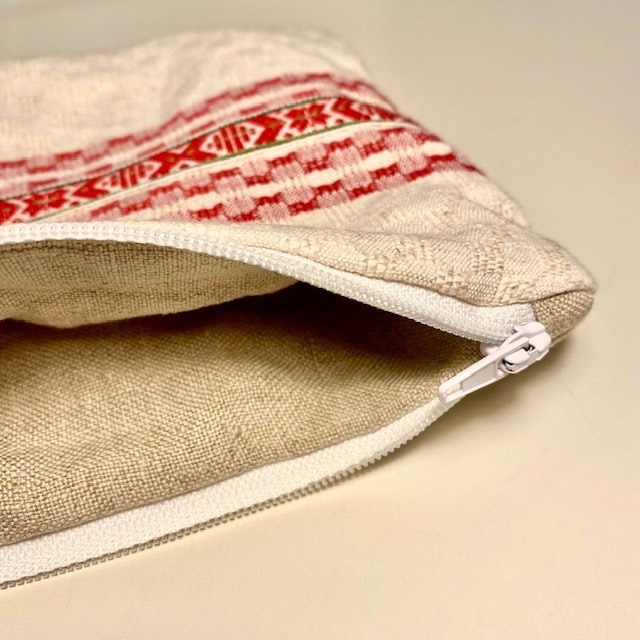 Närbild, miniväska sydd av beige linnetyg och en vit röd kökshandduk. Ett dalaband/hemslöjdsband i röd och vitt är fastsytt som dekoration.