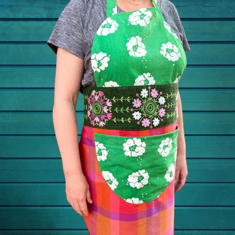 Närbild av förkläde Vimla sytt i grönt retrotyg, med midjebälte av duk med 50-tals broderier i rosa och grönt. Förklädet har ett rundat liv med insnitt och stor magficka.