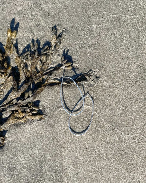 Handtillverkade tunna, ovala örhängen i silver. På bilden ligger örhängena på stranden.