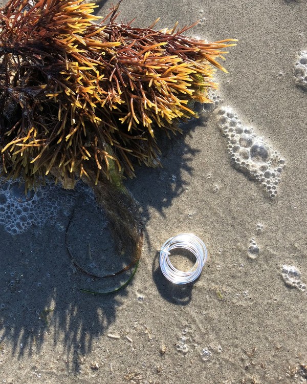 Handgjord ring med flera tunna silverringar, ihopsatta med en liten silverögla. Ligger på stranden med tång.