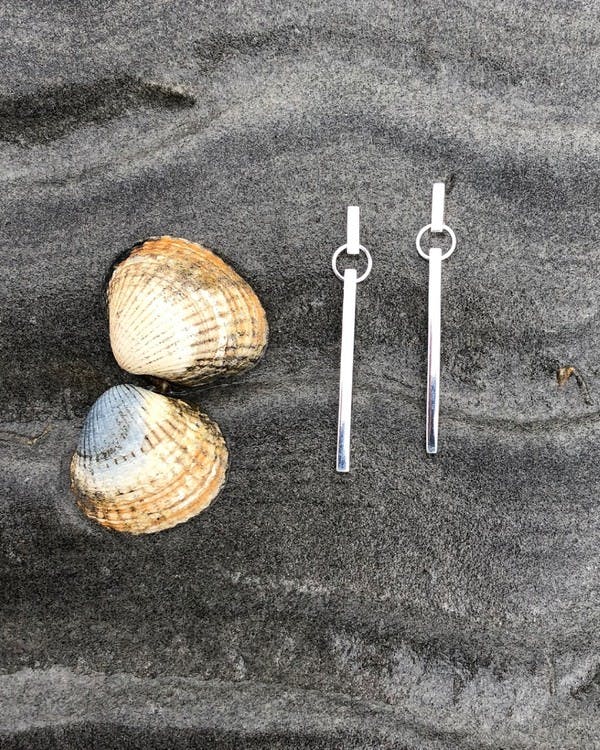 Handgjorda örhängen i silver, gjort av två fyrkantiga pinnar med en ring mellan. Bilden visar två örhängen på stranden.