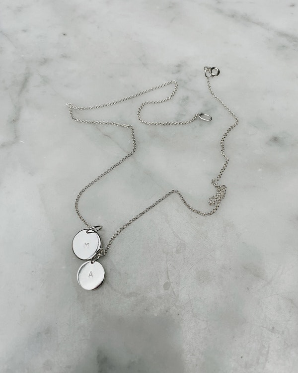 Halsband med bokstavsplatta, med en stämplad bokstav. Handgjort i silver. Bilden visar ett par plattor på en kedja.