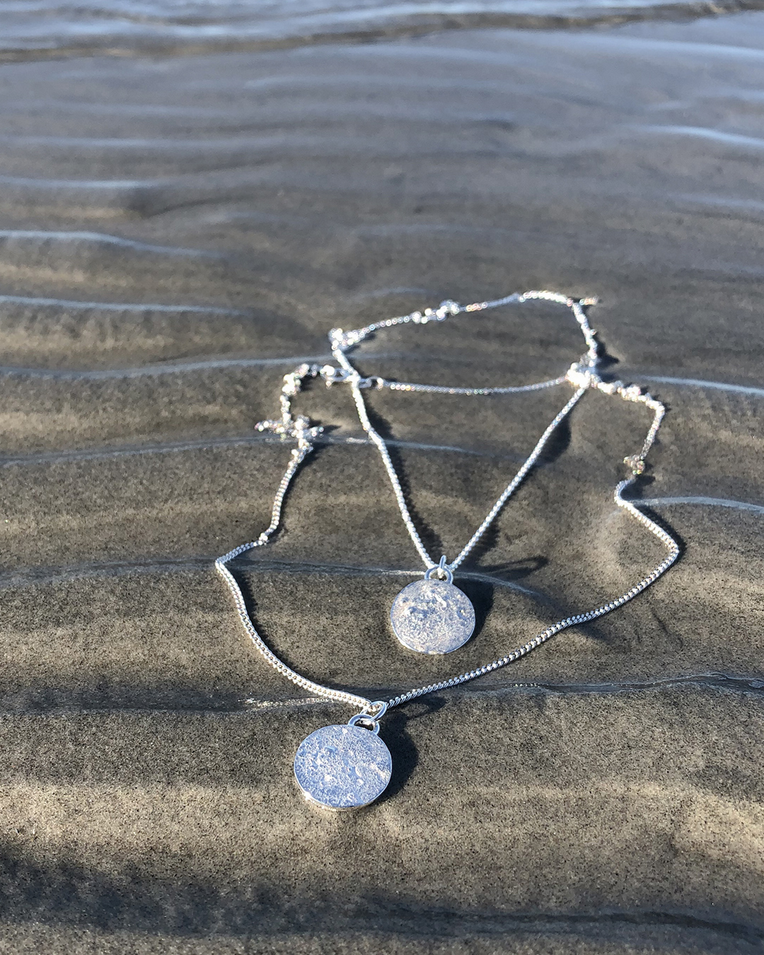 Liten fullmåne i silver, som halsband. Två stycken visas på en strand.