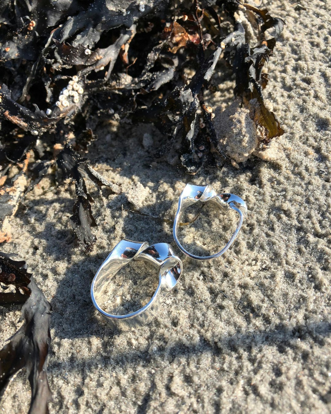 Ring i silver med vriden twist framtill. Två ringar ligger på stranden.