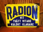 Plan och rektangulär skylt i emaljerad plåt med vikt kant. Motivet är blå och vit text ”Radion, vit tvätt vitare, kulört klarare” mot gul bakgrund med blått fält. Måtten är 350 x 250 mm. Skylten tillv