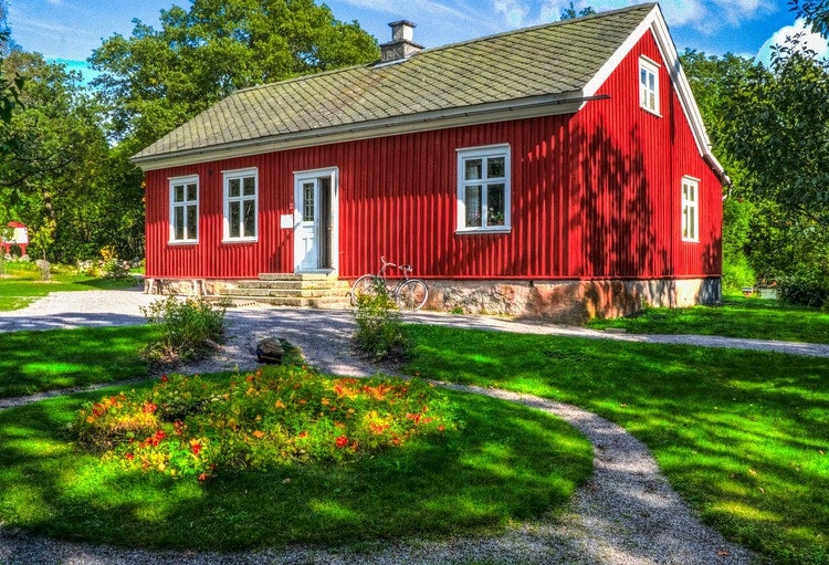 Rött stort äldre hus med gräsmatta framför illustrerar ianspråktagen mark vid strandskydd. Billigare än advokat