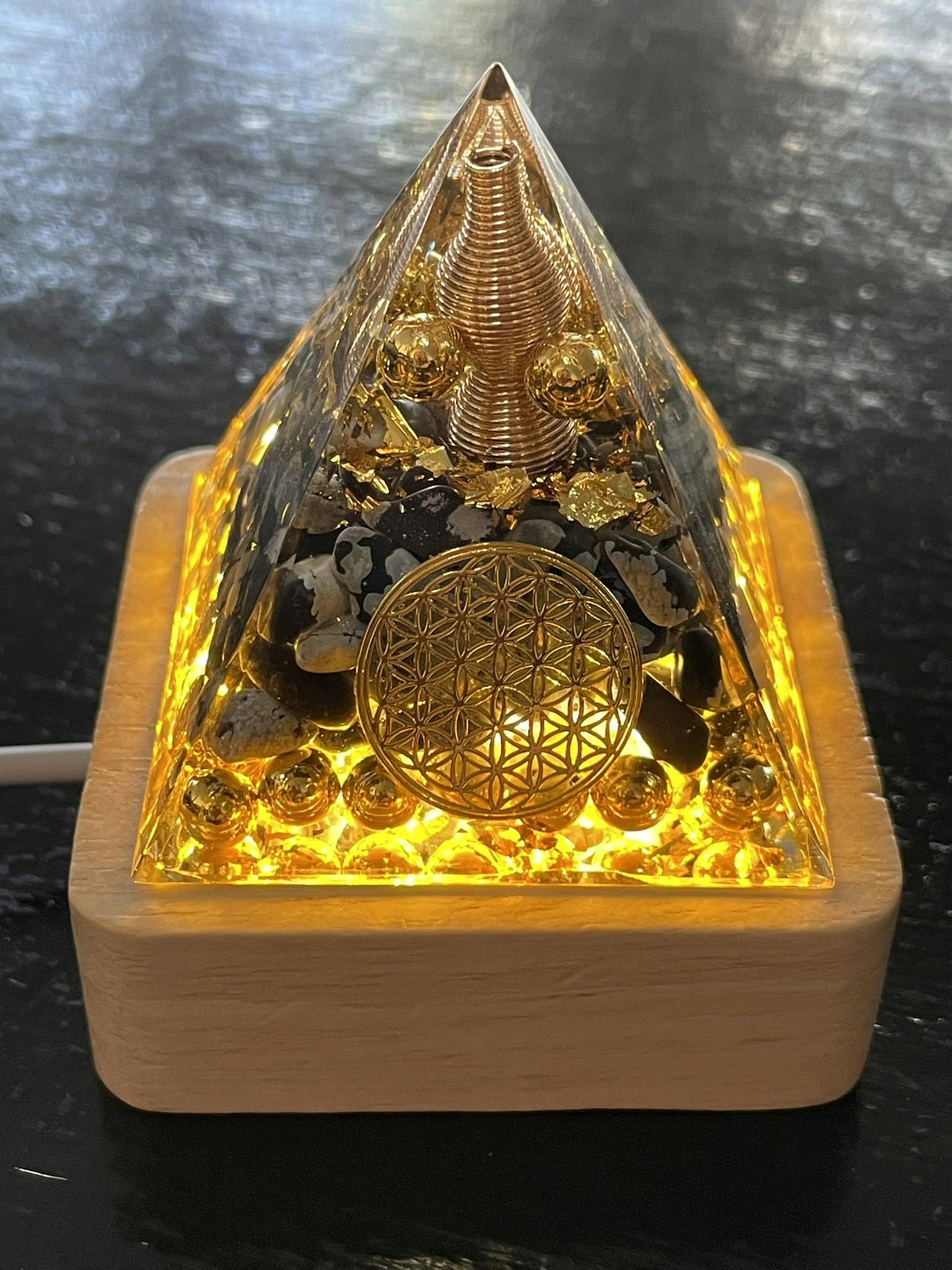 Bilden visar en orgonitpyramid med livetsblomma och metalldetaljer stående på en upplyst underdel.