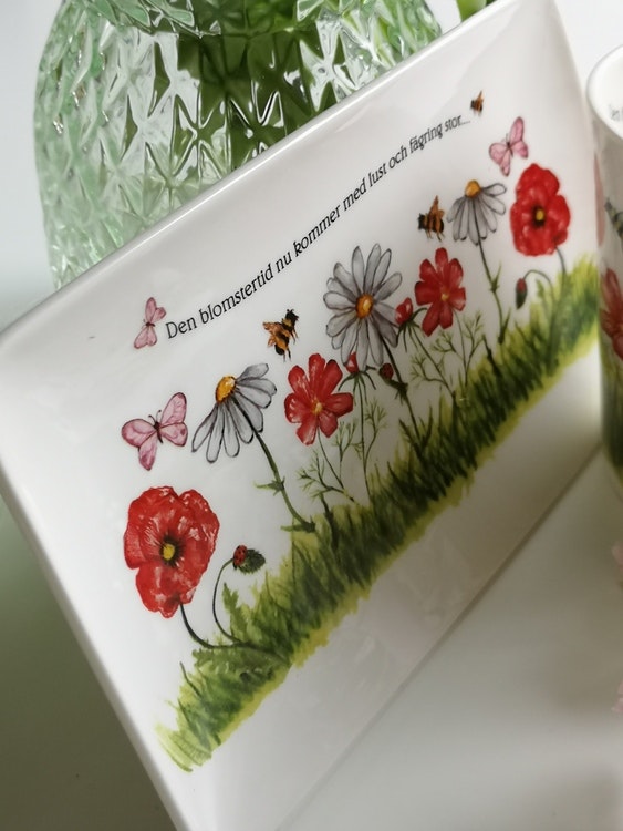 Assiett med rektangulär form med motiv av vita och röda blommor samt fjärilar och bin. Även text på assietten.