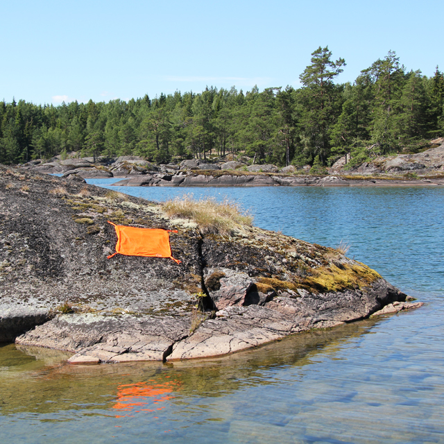 Some distance away shows the Orange Signal Panel placed on rocky ground near water in sunny summer Sweden.