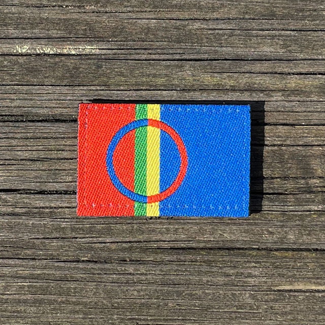 A Sámi Flag Hook Patch Small from TAC-UP GEAR laying flat on a wooded floor