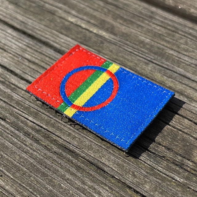 A Sámi Flag Hook Patch Small from TAC-UP GEAR laying flat on a wooded floor seen from an angle