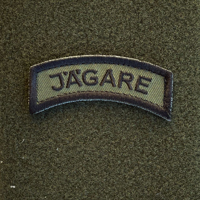 A JÄGARE K3 Type Hook Patch Black/Green seen from the front mounted on a green loop piece on wooded floor