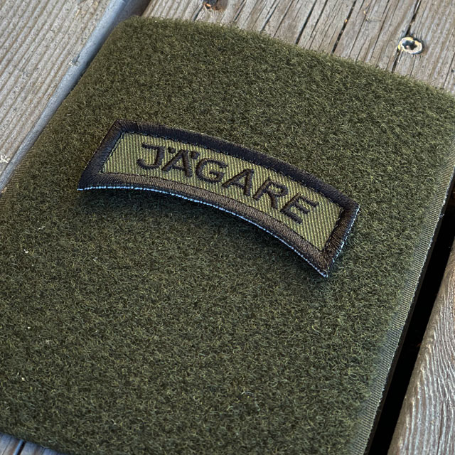 A JÄGARE K3 Type Hook Patch Black/Green seen from an angle mounted on a green loop piece on wooded floor