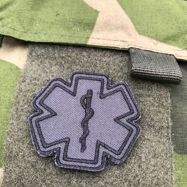 A mounted MEDIC Star of Life Black Grey Hook Patch on a armpocket of a M90 Camouflage jacket.