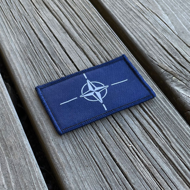 Seen from an angle a NATO Flag Hook Patch from TAC-UP GEAR on a wooded floor