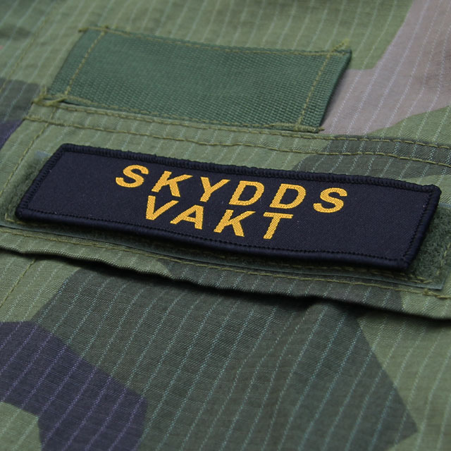 Skyddsvakt Avlång Kardborremärke med M90 kamouflage i bakgrunden.