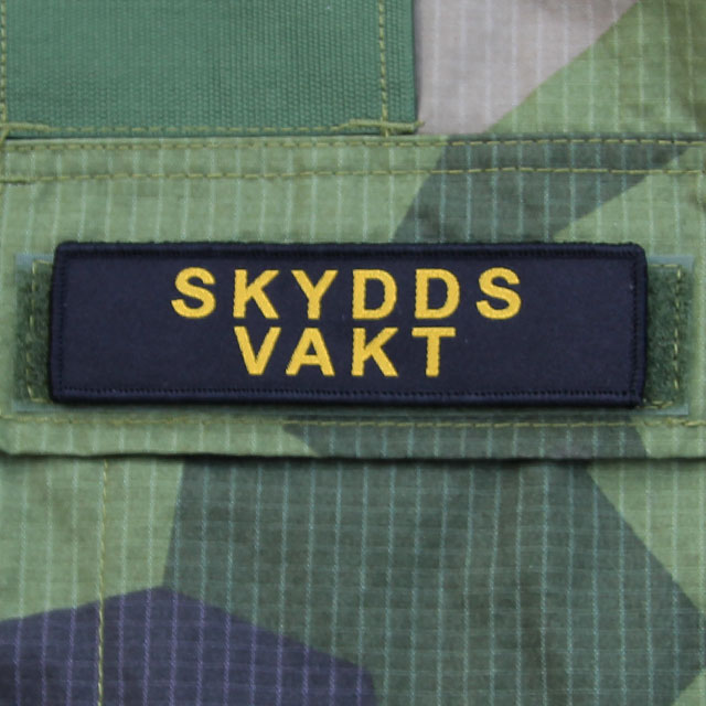 Ett Skyddsvakt Avlång Kardborremärke monterat på locket på en bröstficka på en M90 kamouflage jacka.