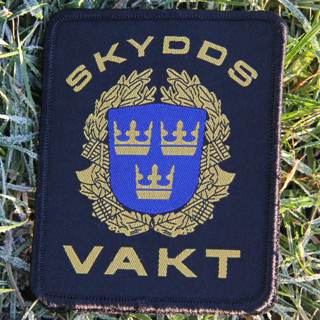 Det militära Skyddsvakt tygmärke kardborre Svart har text och mäke i fint gult och blått mot svart botten.