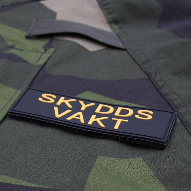 Produktfoto av ett Skyddsvakt PVC Avlång Kardborremärke mot en bakgrund av M90 kamouflage.