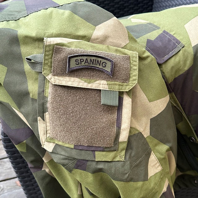 A SPANING Tab Hook Patch Black/Green from TAC-UP GEAR seen stright on and mounted on an arm pocket on a NCWR Jacket M90