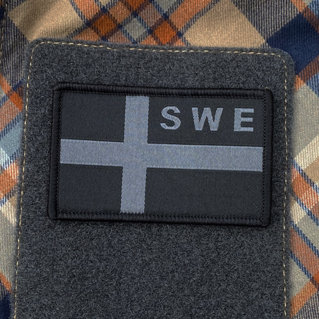 A Sweden Flag OPS Nylon Black/Grey Patch mounted with velcro on an plaid shirt